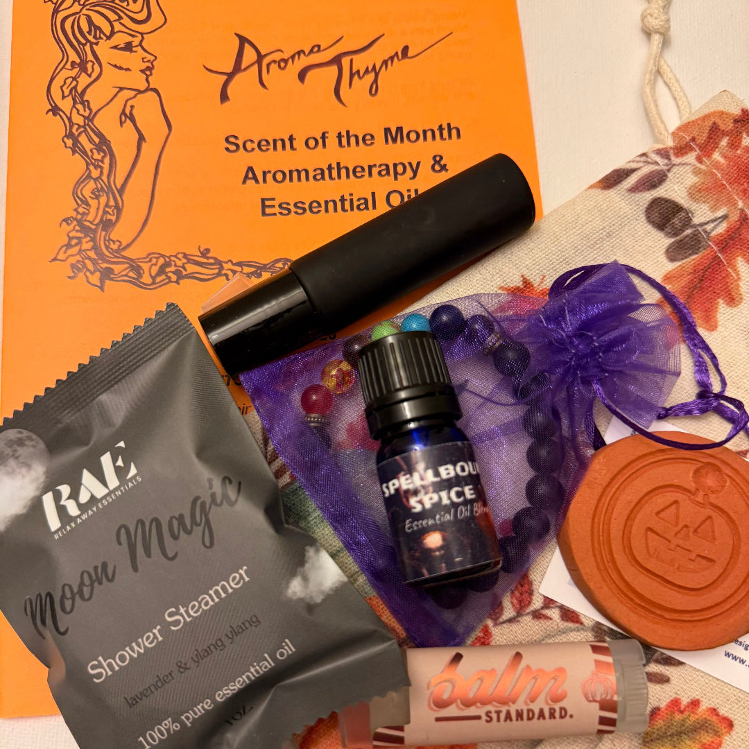 A curated autumn-themed aromatherapy and essential oil gift set arranged on a white surface. The set includes an orange Aroma Thyme 'Scent of the Month' card, a black roller bottle, a gray RAE 'Moon Magic' shower steamer packet, a small blue 'Spellbound Spice' essential oil bottle, a terracotta pumpkin diffuser, a purple chakra beaded bracelet in a sheer purple bag, and a pumpkin spice Balm Standard lip balm.