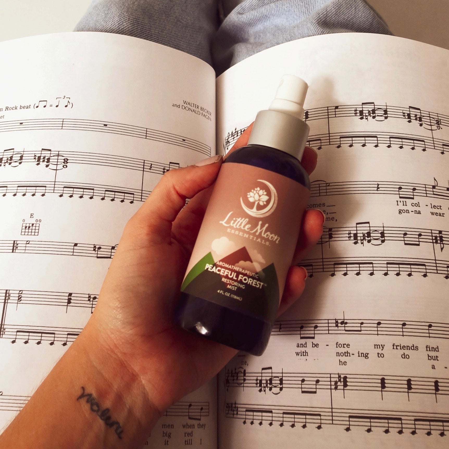 Hand holding a bottle of 'Little Moon' Peaceful Forest Spray Mist product on sheet music
